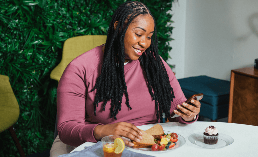 African-American woman smiles at her phone while eating a sandwich