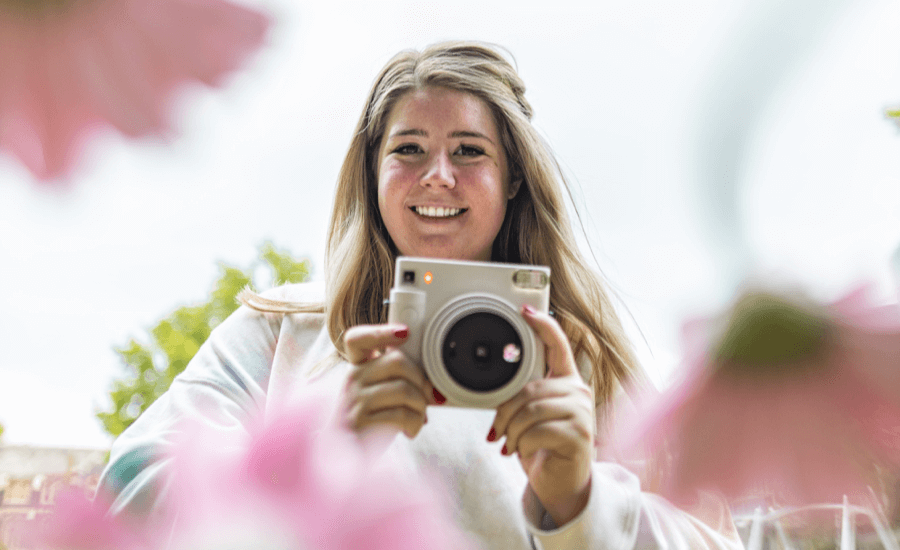 Blonde woman holds a white camera with blurry flowers in the foreground