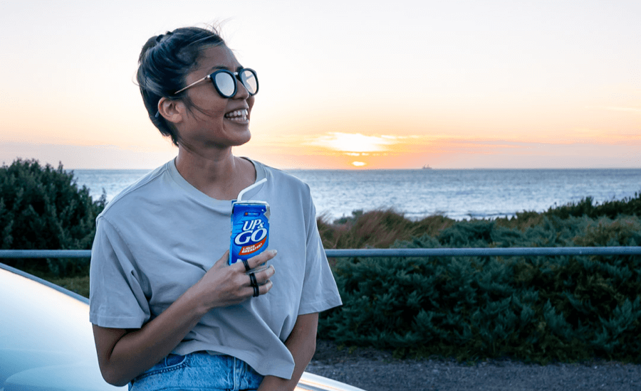 Woman with sunglasses sits on bonnet of car drinking an Up & Go at sunrise