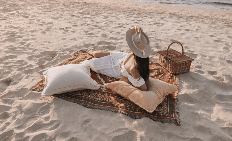 Woman with a broad brimmed hat sits on a rug with pillows on a beach at sunset.