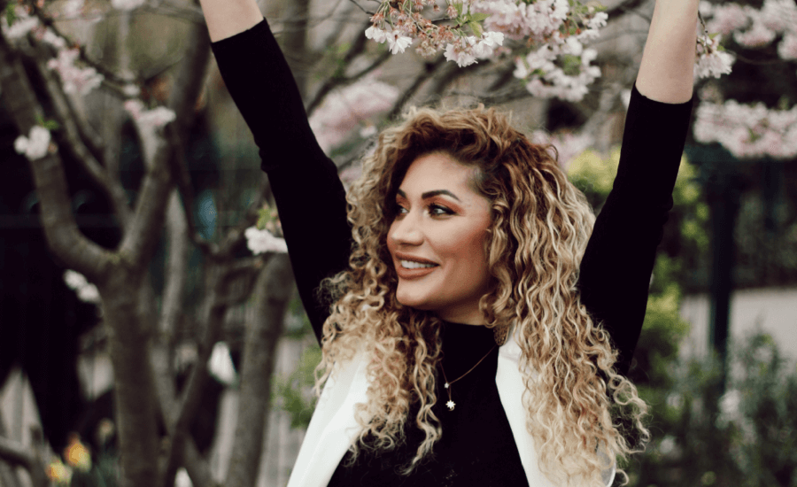 Woman sitting under a cherry blossom tree looking to the right with her arms up in the air