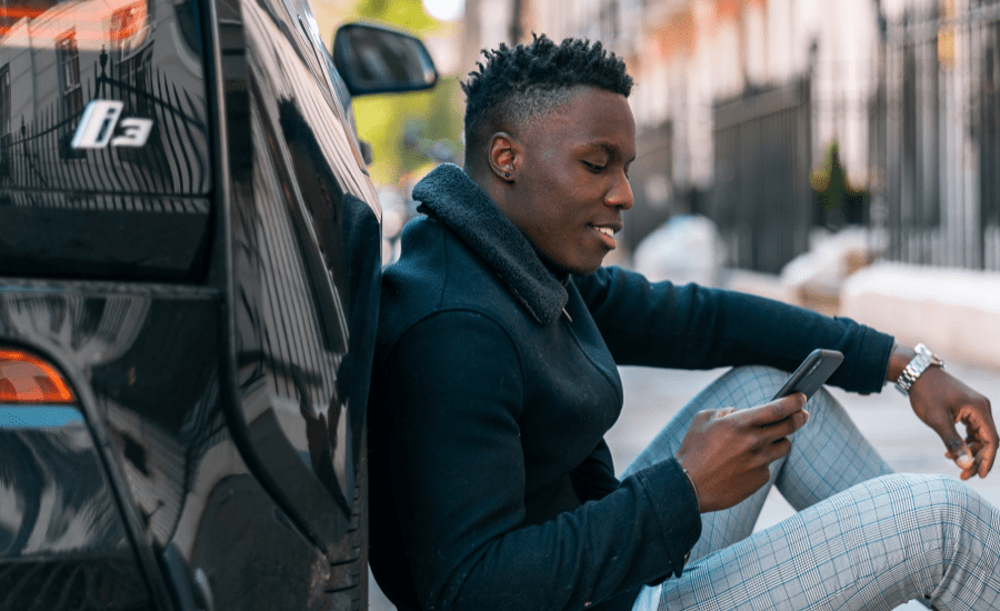 Man looks at his phone while leaning seated against a car