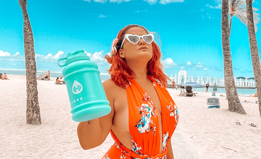 Woman in swimwear and sunglasses holds up water bottle at the beach