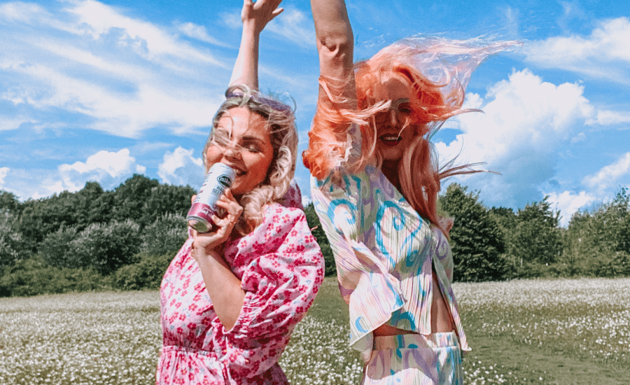 two women out in a field facing the camera and smiling while holding a can, hair blowing in the wind