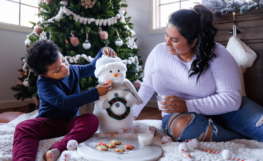 mother and child sit in front of a christmas tree decorating a snowman