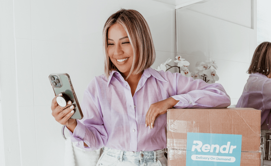 Woman smiles while looking at phone with an arm leaning against a cardboard box