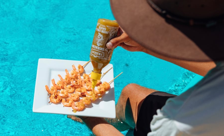 TRIBE creator @unchartedaus pouring sriracha sauce onto a plate of food by the pool for Trident